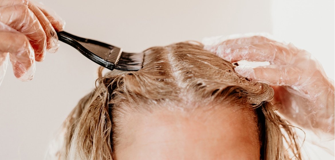 Ein blonder Frauenscheitel von vorne. Die Frau trägt durchsichtige Handschuhe und appliziert mit einem schwarzen Pinsel Blondierung auf ihren Haaransatz.